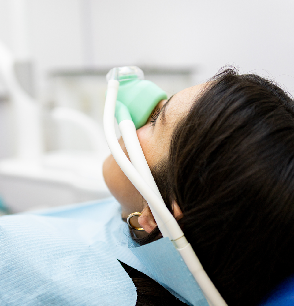 An adult woman sits in a dentist's clinic wearing a nasal mask to inhale nitrous oxide